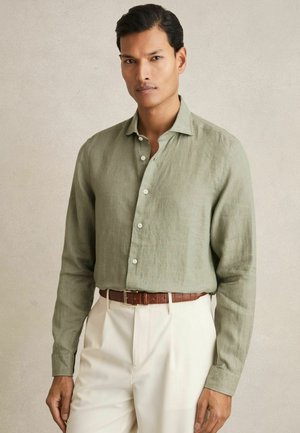 Une chemise en lin vert clair à manches longues avec un col boutonné, un devant à patte de boutonnage et assortie à un pantalon crème et une ceinture marron.