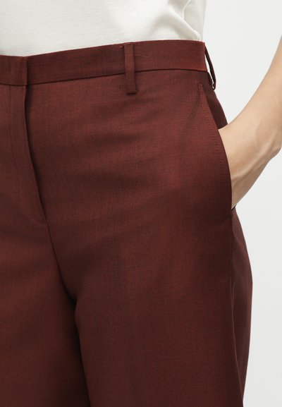 Άτομο που φοράει σκούρο κόκκινο παντελόνι ραμμένο στα μέτρα του, με το δεξί του χέρι στην πλαϊνή τσέπη, σε συνδυασμό με λευκό τοπ.