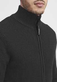 Svart zip-up genser med en ribbet, teksturert stoff. Har høy krage og en merket glidelås, egnet for uformell bruk.