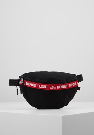 Sac banane noir avec une bande de fermeture éclair rouge affichant "REMOVE BEFORE FLIGHT" en lettres blanches, présenté sur un piédestal blanc.