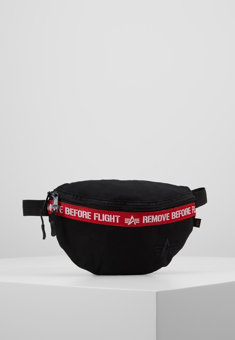 Sac banane noir avec une bande de fermeture éclair rouge affichant "REMOVE BEFORE FLIGHT" en lettres blanches, présenté sur un piédestal blanc.