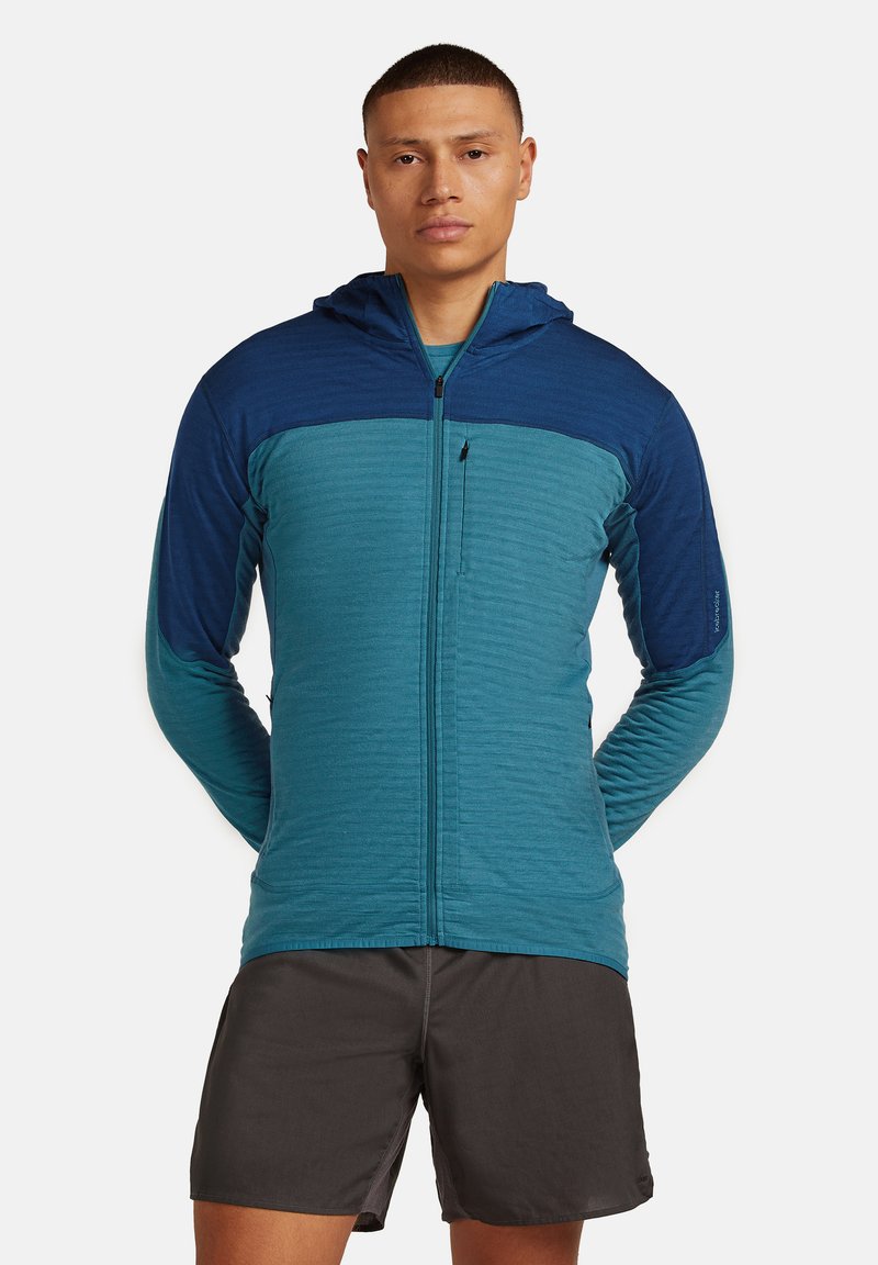 Uomo che indossa una felpa blu con zip e cappuccio, insieme a pantaloni corti grigio scuro, in piedi con le mani dietro la schiena su uno sfondo bianco.