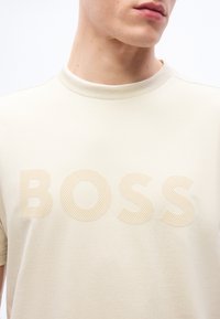 Κοντινό πλάνο ενός ατόμου που φοράει μπεζ μπλουζάκι με ανάγλυφη επιγραφή «BOSS» στο στήθος και ριμπ γιακά.