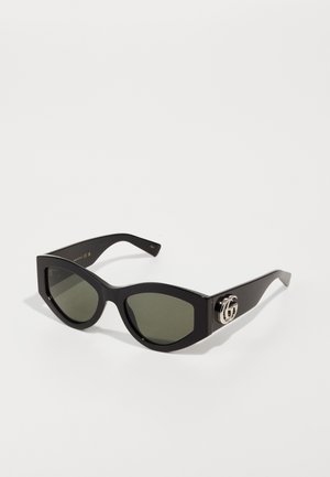 Gafas de sol negras en forma de ojo de gato con lentes verdes, acabado brillante y detalles del logo en plata en la patilla. Montura angular con brazos gruesos.
