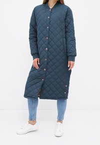 Femme portant un long manteau matelassé bleu sarcelle foncé avec des boutons-pression, associé à un jean bleu clair et des baskets blanches, debout devant un fond blanc.