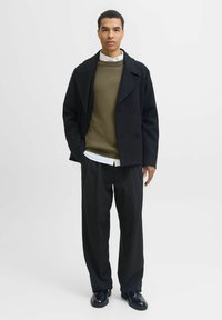 Jauns vīrietis stāv, ģērbies melnā pīlera mētelī, olīvzaļā džemperī pāri baltajai krekla, melnās svītrainās biksēs un melnās uzvalka kurpēs uz viendabīga fona.