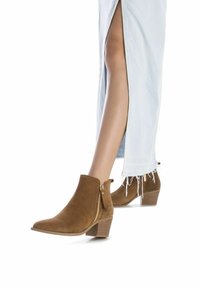 Botines de ante marrón con cremallera lateral, detalles texturizados y tacón apilado, exhibidos junto a una falda de mezclilla clara con flecos.