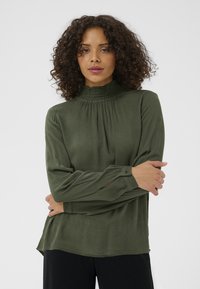 Blouse verte à manches longues avec un col montant smocké, tissu texturé, coupe ample et poignets boutonnés ; design à plusieurs couches avec un ourlet arrondi.