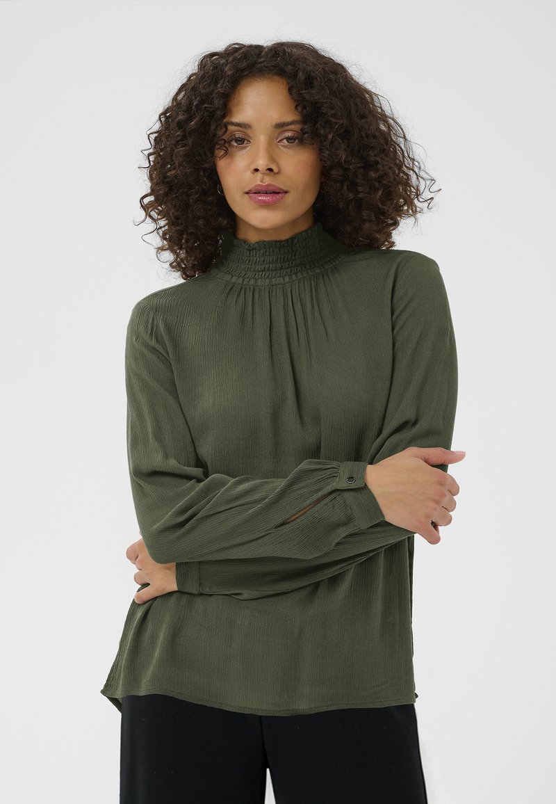 Blouse verte à manches longues avec un col montant smocké, tissu texturé, coupe ample et poignets boutonnés ; design à plusieurs couches avec un ourlet arrondi.