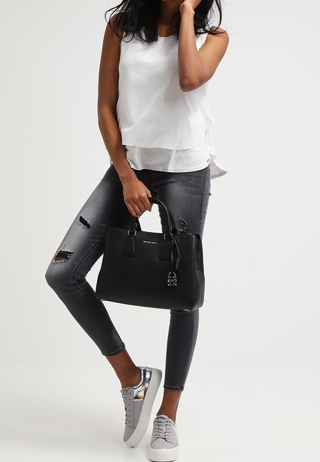 Femme portant un haut blanc sans manches et un jean noir déchiré, tenant un sac à main noir, debout, une jambe croisée, portant des baskets grises.