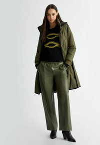 Cappotto puffo verde, maglione nero con motivo giallo, pantaloni in pelle verde, stivaletti neri con tacco; texture lisce e vestibilità moderna.