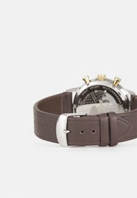 Montre avec bracelet en cuir marron, au dos en acier inoxydable et aux accents dorés. Elle dispose d'un cadran rond, de sous-cadrans chronographes et d'une boucle en argent.