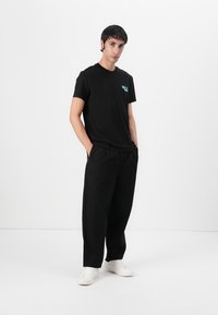 Black cotton t-shirt with a turquoise graphic on the chest, paired with black wide-leg trousers and white sneakers, set against a plain background.