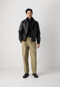 Čierna kožená bunda cez čierny sveter so zipsom, doplnená khaki cargo nohavicami a čiernymi topánkami. Tento look sa vyznačuje vreckami a voľným strihom.