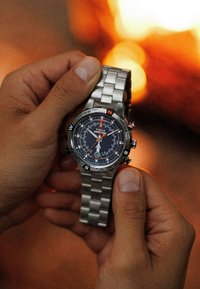 Montre en acier inoxydable argenté avec un cadran bleu, des accents orange et une lunette de compas. Elle possède un bracelet métallique et est tenue entre deux mains.