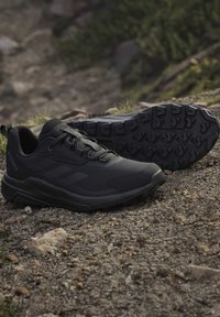Zwarte trailrunningschoenen met textuurrijke zolen geplaatst op rotsachtige, modderige grond buiten.