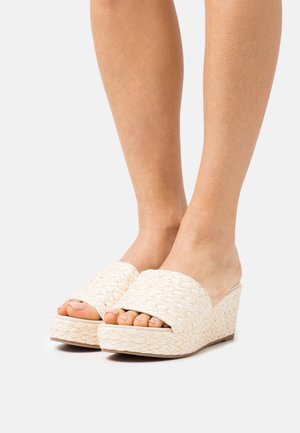 Pieds portant des sandales compensées en toile beige avec orteils ouverts sur un fond blanc.