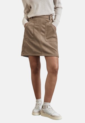 Mini-jupe en suède marron avec passants de ceinture, poches avant et coutures plates. Associée à des baskets blanches et beiges et portée avec un léger pull.