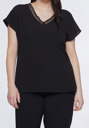 Kobieta ubrana w czarną bluzkę z dekoltem w serek wykończoną koronką oraz dopasowane czarne spodnie, stojąca na jednolitym jasnym tle.