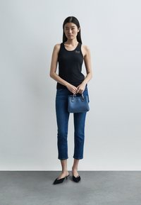 Γυναίκα με μαύρο τοπ χωρίς μανίκια, κοντό τζιν, μαύρες γόβες, κρατώντας μικρή μπλε τσάντα, στέκεται μπροστά σε απλό ανοιχτό γκρίζο φόντο.