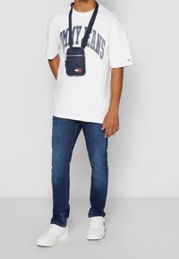 Vit t-shirt med tryck av "TOMMY JEANS", mörkblå axelväska, blå jeans och vita sneakers. Avslappnad outfit med modern design.