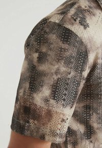 Chemise à manches courtes en tissu transparent avec une texture semblable à de la dentelle dans des nuances de brun et de beige, présentant des motifs floraux et géométriques.