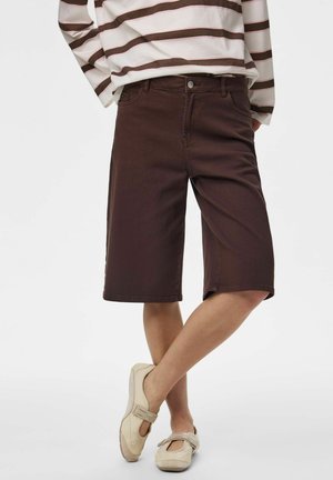 Personne portant un short marron foncé jusqu'aux genoux, un haut à manches longues blanc et marron rayé et des chaussures beiges, debout jambes croisées.
