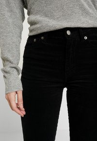 Svarta kordflöjlsbyxor med vertikal ribbad textur, med hög midja, silverdetaljer och en grå stickad tröja som är delvis synlig.