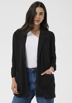 Kvinde iført en sort blazer over en hvid ribbet top og blå jeans, med hånden i blazerlommen, stående mod en ensfarvet baggrund.