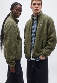 To personer iført olivengrønne fleecejakker med sidelommer med lynlås, tekstureret stof og høje kraver, stående mod en ensfarvet baggrund.