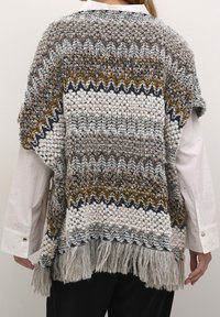 Kvinna som bär en texturerad stickad poncho med zigzagmönster och fransig fåll över en vit långärmad skjorta, sedd bakifrån.