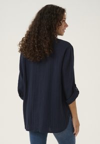 Camicia a maniche lunghe blu navy con una finitura testurizzata, maniche arrotolate e orlo curvo, abbinata a jeans azzurri.