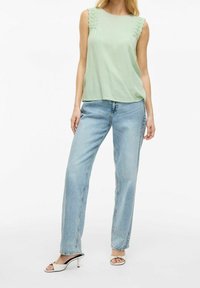 Jasnozielony top bez rękawów z teksturowanymi akcentami na ramionach, w połączeniu z jasnoniebieskimi jeansami o prostych nogawkach i białymi sandałami na obcasie z odkrytymi palcami.