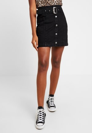 Femme portant une mini-jupe noire boutonnée devant avec ceinture, un haut imprimé léopard et des baskets montantes noires avec lacets blancs.