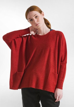 Maglione rosso oversize con scollatura a barco, maniche lunghe e due tasche frontali. Realizzato in tessuto morbido e strutturato. Pantaloni neri parzialmente visibili.