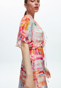 Robe maxi colorée et transparent présentant un motif géométrique abstrait en rose, orange et bleu. Une taille nouée et des manches volantées ajoutent des détails.