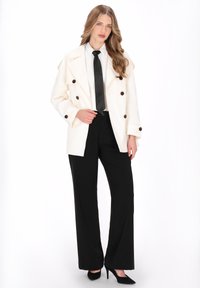 Fehér kétsoros blézer barna gombokkal, fehér inggel, fekete nyakkendővel és fekete, széles szárú nadrággal párosítva. A megjelenést fekete magassarkú cipő teszi teljessé.