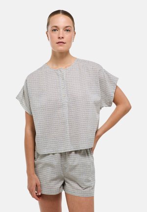 Femme aux cheveux attachés portant un haut à manches courtes à carreaux boutonné et un short assorti, debout avec une main sur la hanche.