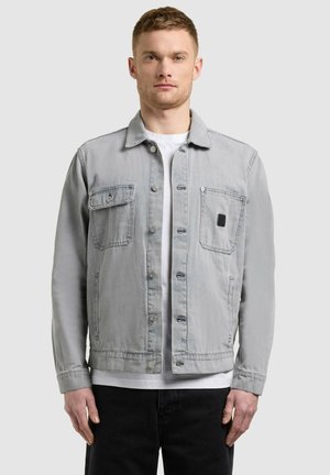 Muž v svetlosivej zapínanej bunde s dvoma náprsnými vreckami, bielou košeľou a čiernymi nohavicami, stojaci pred jednoliatym pozadím.