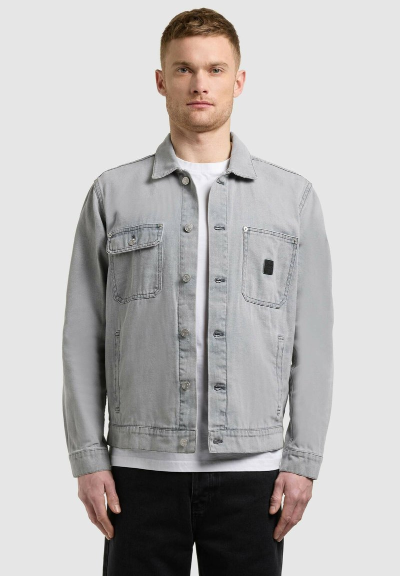 Homme portant une veste boutonnée gris clair avec deux poches poitrine, sur une chemise blanche et un pantalon noir, debout devant un fond uni.