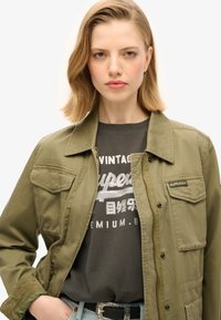 Olivengrøn arbejdsjakke med to brystlommer og trykknapper, lagdelt over en mørk grafisk t-shirt med sølvtekst. Slidte denimjeans ses.