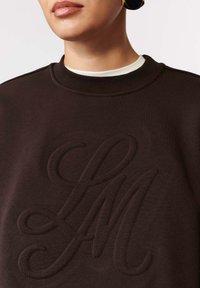 Zbliżenie na osobę w ciemnobrązowej bluzie z podniesionymi, kursywą napisanymi inicjałami "LM" oraz białej koszuli pod spodem.