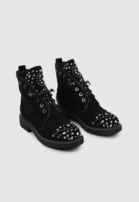 Botas de tobillo de ante negro con puntera redonda y suela plana, adornadas con acentos de tachuelas plateadas y aqua, así como ojetes decorativos en el cañón.