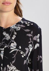 Blommig blus med en mörk bakgrund och blommönster i ljusgrått och vitt, knäppning fram och ett mjukt, texturerat tyg.