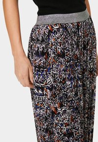 Flerfärgad, mönstrad plisserad kjol med ett silverfärgat elastiskt midjeband. De dominerande färgerna inkluderar lila, orange, blå och svart.