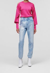 Eine Frau mit einem rosa Oberteil mit kalten Schultern, hoch taillierten hellblauen Jeans mit weißen Sprenkeln und weißen Stiefeletten, die vor einem weißen Hintergrund steht.