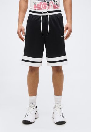 Άνδρας που φοράει μαύρο και λευκό σορτς μπάσκετ με κορδόνι και λευκά αθλητικά παπούτσια με μαύρες λεπτομέρειες, στέκεται σε λευκό φόντο.