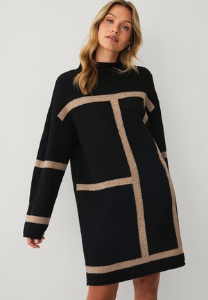 Kvinde med langt blondt hår iført en løs, sort sweaterkjole med beige geometriske linjer, stående foran en ensartet lys grå baggrund.