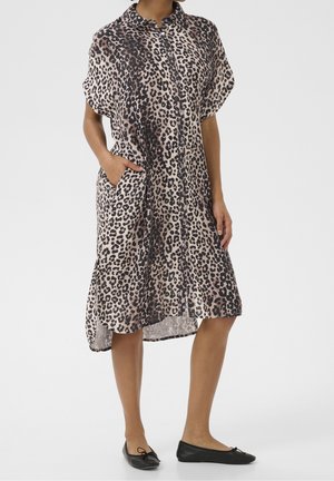 Kobieta ubrana w luźną midi sukienkę na guziki w panterkę z kieszeniami, zestawioną z czarnymi balerinami na białym tle.