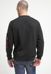 Sudadera negra con cuello redondo, tela texturizada y puños de canalé. Cuenta con un bolsillo con cremallera en el brazo izquierdo, combinada con vaqueros azules.
